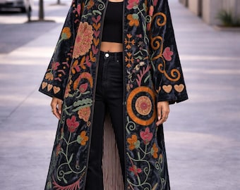 May include: A long, dark blue velvet coat with colorful floral embroidery. The coat features a mandarin collar and a long, flowing design. The embroidery includes orange, pink, and green flowers and leaves. The coat is worn over black pants and a black top.