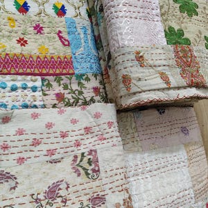 May include: A stack of colorful, embroidered quilts. The quilts feature a patchwork design with various floral and geometric patterns in shades of cream, pink, blue, green, and yellow. The stitching is visible, adding texture and detail to the textiles.