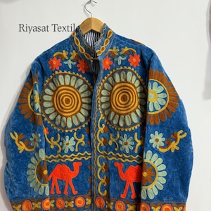 May include: A blue embroidered jacket with a mandarin collar. The jacket features orange and yellow floral patterns, green and yellow circular designs, and two orange camel figures. The text "Riyasat Textile" is visible on the upper left.