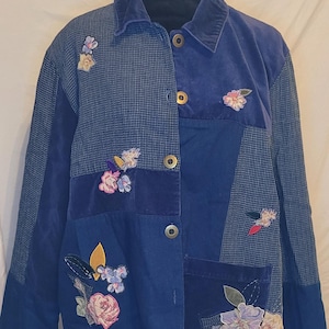 May include: A blue patchwork jacket with floral embroidery. The jacket features a corduroy collar, button closures, and a mix of solid and patterned fabrics. Appliqued flowers in pink, white, and yellow add a decorative touch to the garment.