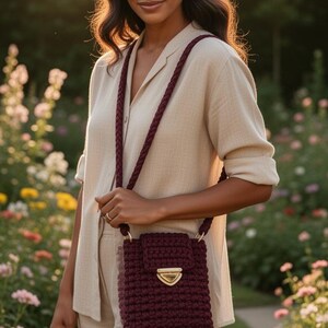 May include: A burgundy crochet crossbody bag with a gold-colored clasp. The bag has a braided strap and is worn over the shoulder. The person in the image is wearing a beige outfit and standing in a garden.