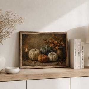 May include: A rectangular framed wall art print depicting a still life scene with four pumpkins in shades of white, green, and orange, a lit candle in a dark holder, and a vase filled with dried autumn leaves on a textured surface.