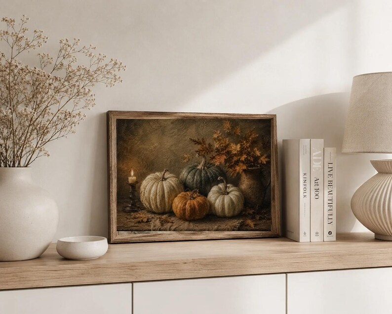 May include: A rectangular framed wall art print depicting a still life scene with four pumpkins in shades of white, green, and orange, a lit candle in a dark holder, and a vase filled with dried autumn leaves on a textured surface.