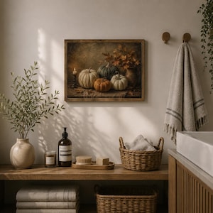 May include: A rectangular framed wall art print featuring a still life painting of white, green, and orange pumpkins, autumn leaves, and a lit candle. The artwork is displayed in a bathroom setting with a wooden shelf, wicker baskets, and towels.