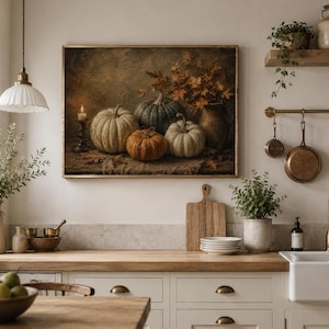 May include: A framed wall art print depicting a still life of four pumpkins in shades of white, green, and orange, arranged with a lit candle and autumn foliage in a vase. The artwork features a dark, textured background and sits above a kitchen counter.