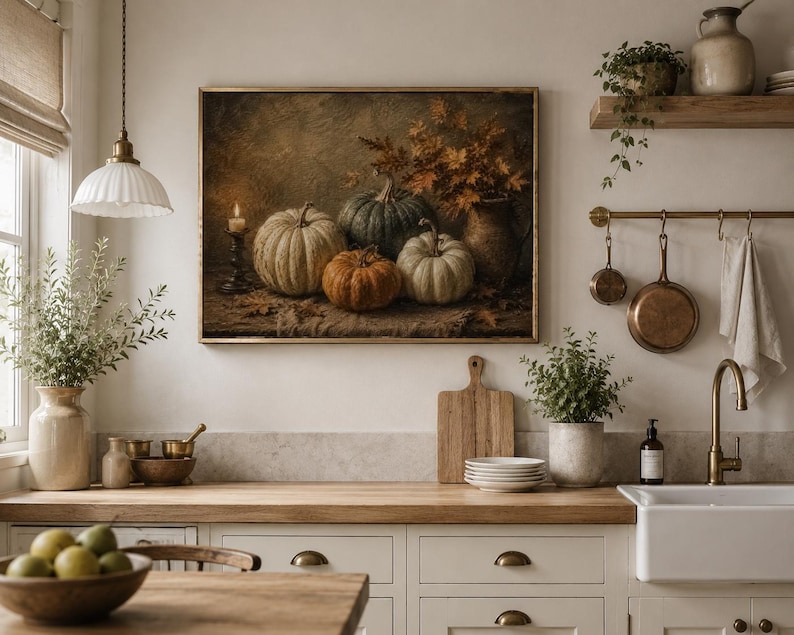 May include: A framed wall art print depicting a still life of four pumpkins in shades of white, green, and orange, arranged with a lit candle and autumn foliage in a vase. The artwork features a dark, textured background and sits above a kitchen counter.