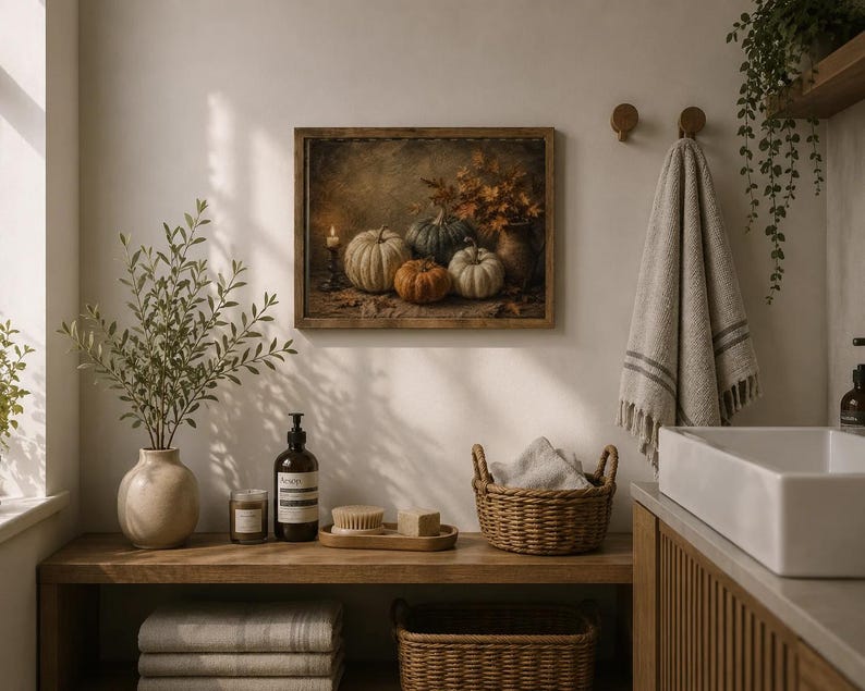 May include: A rectangular framed wall art print depicting a still life of pumpkins, a lit candle, and autumn leaves in muted tones. The artwork is displayed on a light-colored wall above a wooden vanity featuring a plant, soap, and baskets.