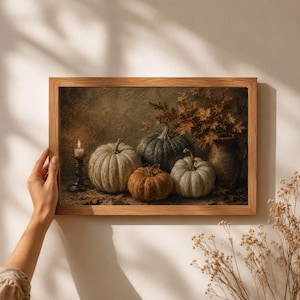 May include: A framed wall art print featuring a still life composition of three pumpkins in shades of green, white, and orange, a lit candle in a dark metal holder, and a rustic ceramic pitcher filled with dried autumn leaves on a textured surface.
