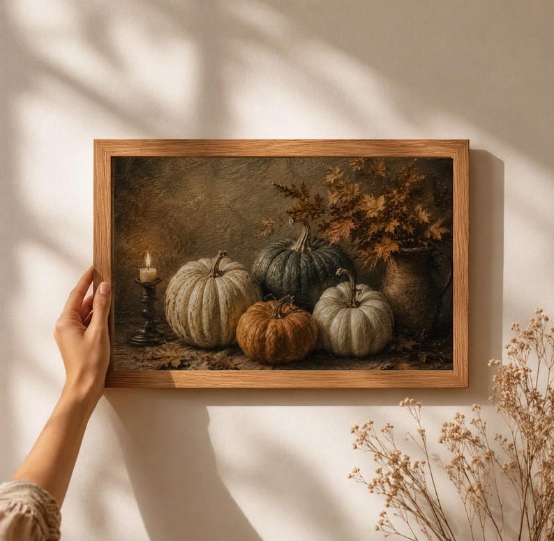 May include: A framed wall art print featuring a still life composition of three pumpkins in shades of green, white, and orange, a lit candle in a dark metal holder, and a rustic ceramic pitcher filled with dried autumn leaves on a textured surface.
