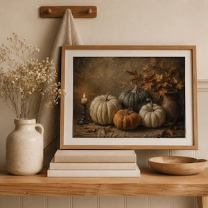 May include: A framed wall art print depicting a still life of four pumpkins in shades of green, white, and orange, a lit candle in a dark holder, and a vase with autumn leaves, all arranged on a textured surface with scattered dried leaves.