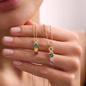 May include: Two gold necklaces with heart-shaped gemstones in various colors. Each necklace features a pendant with an infinity symbol design, followed by three heart-shaped gems. The gemstones are green, yellow, and red on one necklace, and green, pink, and purple on the other.