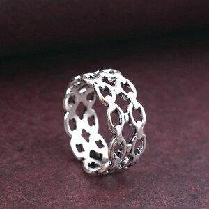 May include: A silver-colored ring with a chain-link design. The ring has an openwork pattern, with each link connected to the next. The ring is made of a shiny metal and is set against a dark red background.