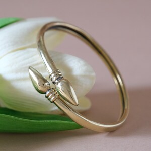May include: A gold-coloured cuff bracelet with a distinctive design. The bracelet has two decorative, pointed ends that meet in the centre. The bracelet is set against a white flower and green leaf, with a soft pink background.