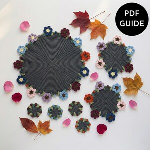May include: A collection of dark gray crochet placemats and coasters, each adorned with colorful crocheted flowers. The image includes fallen leaves and rose petals, with a black circle that says "PDF GUIDE".