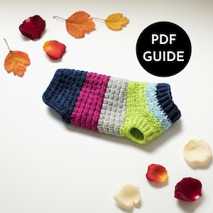 May include: A colorful, striped dog sweater in shades of navy, fuchsia, gray, and lime green. The sweater has a ribbed collar and a hole for the dog's legs. The image includes the text "PDF GUIDE" in a black circle, along with autumn leaves and flower petals.