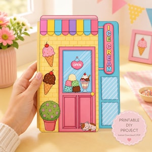 Puede incluir: Una fachada de heladería de papel con una pared de ladrillo amarillo, una puerta rosa con un letrero de abierto y un cartel azul que dice ICE CREAM. El diseño incluye ilustraciones de conos de helado, una planta en maceta y un gato dormido.