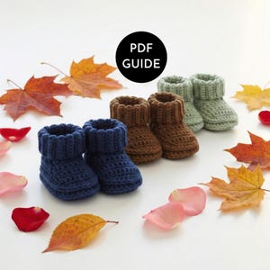 Può includere: Scarpine da neonato all'uncinetto fatte a mano nei toni del blu, marrone e verde. Le scarpine sono disposte con foglie autunnali e petali di rosa, con una grafica "PDF GUIDE".