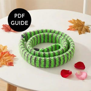 May include: A coiled, green and gray striped crocheted rope. The rope is circular and has a loop at one end. The image includes a black circle with the text "PDF GUIDE". Scattered around the rope are autumn leaves and rose petals.