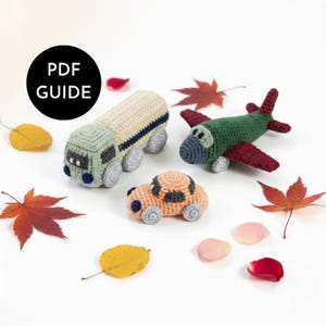 May include: A collection of crocheted toy vehicles, including a truck, car, and airplane, in various colors. The truck is beige and green, the car is peach, and the airplane is green and red. The image includes fall leaves and the text "PDF GUIDE".
