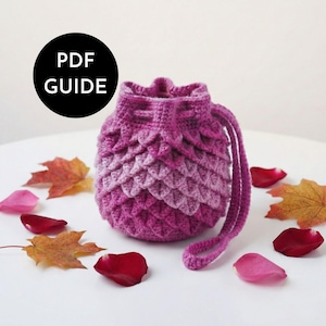 Puede incluir: Un bolso con cordón hecho a mano, tejido a ganchillo en tonos rosas y morados, con un patrón texturizado en forma de escamas. El bolso tiene una correa a juego y está rodeado de hojas de otoño y pétalos de rosa. La imagen incluye un círculo negro con el texto "PDF GUIDE".