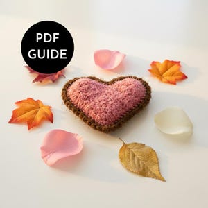 May include: A handmade, pink crocheted heart with a brown border, surrounded by autumn leaves and flower petals. The image includes the text "PDF GUIDE" in a black circle, suggesting a craft tutorial or pattern.