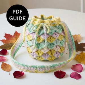 May include: A crocheted drawstring bag in pastel colors, with a scale-like pattern. The bag has a long strap and is surrounded by autumn leaves and rose petals. The text "PDF GUIDE" is in a black circle.