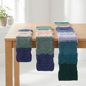 May include: Three hand-knitted, rectangular scarves in varying sizes, arranged on a wooden table. Each scarf features a striped pattern with shades of blue, green, and pink. The largest scarf is approximately 60 inches long, while the smallest is about 20 inches.