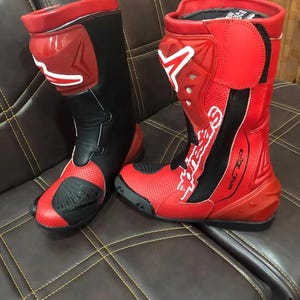 Puede incluir: Par de botas de moto rojas y negras. Las botas tienen un diseño de caña alta con elementos de protección. Las botas tienen el nombre de la marca "Alpinestars" en letras blancas. Las botas son de cuero y tienen un diseño perforado en la puntera.