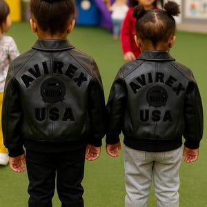 May include: Two children wearing black leather jackets with the text "AVIREX USA" on the back. One child wears black pants, the other gray pants and sneakers. The jackets have a classic bomber style.