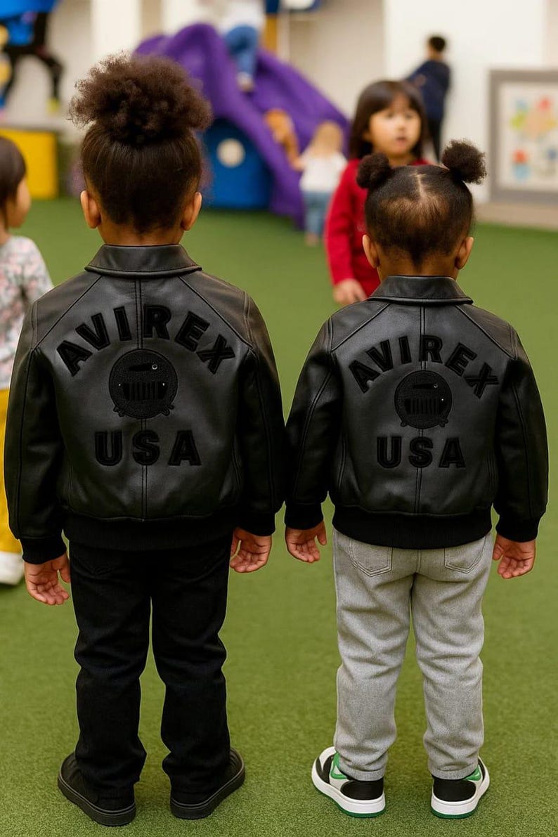 May include: Two children wearing black leather jackets with the text "AVIREX USA" on the back. One child wears black pants, the other gray pants and sneakers. The jackets have a classic bomber style.