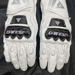 May include: Pair of white motorcycle gloves with black and silver accents. The gloves feature the brand name "DAINESE" and a logo on the back of the hand. The gloves have protective padding on the knuckles and fingers.