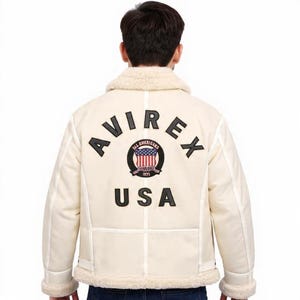 May include: Cream-coloured jacket with a shearling collar and trim. The back features the word "AVIREX" in black block letters above a circular emblem with the American flag and the word "USA" below. The jacket has a classic, vintage style.