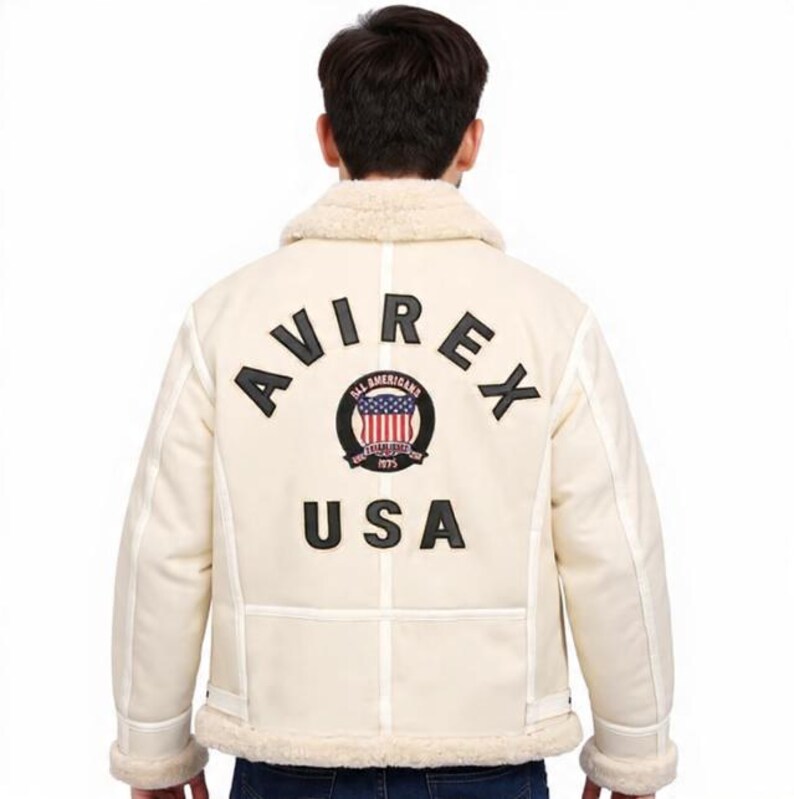 May include: Cream-coloured jacket with a shearling collar and trim. The back features the word "AVIREX" in black block letters above a circular emblem with the American flag and the word "USA" below. The jacket has a classic, vintage style.