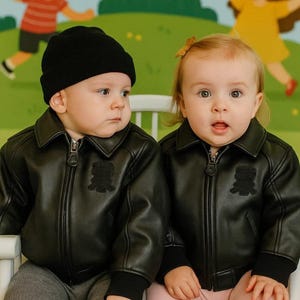 May include: Two babies wearing black leather jackets with a teddy bear patch, black beanies, and gray and pink leggings. They are sitting on a white chair in front of a colorful mural.