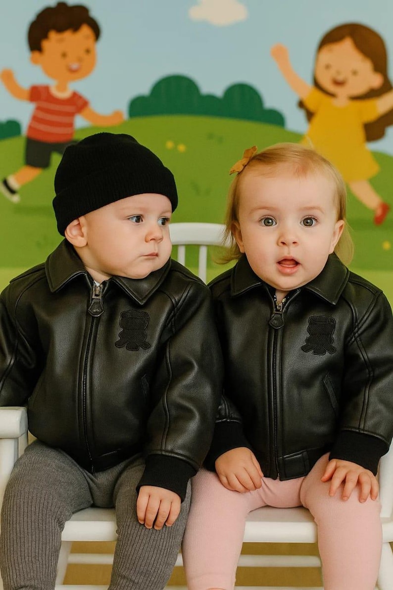 May include: Two babies wearing black leather jackets with a teddy bear patch, black beanies, and gray and pink leggings. They are sitting on a white chair in front of a colorful mural.