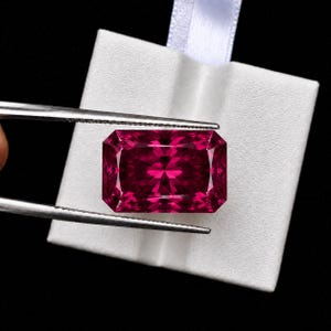 May include: A rectangular, faceted gemstone with a deep, vibrant magenta-red colour. The stone is held by silver-toned metal tweezers against a white, square-shaped display card that features a small white ribbon loop at the top edge.