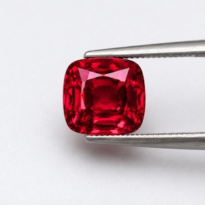Könnte beinhalten: Ein leuchtend tiefroter Edelstein im Kissenschliff mit zahlreichen Facetten, die das Licht reflektieren. Der Edelstein wird von einer silberfarbenen Metallpinzette gehalten und zeigt seine intensive Farbe sowie die symmetrische, abgerundete quadratische Form.