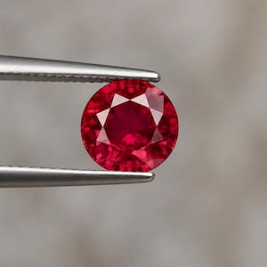 Puede incluir: Una piedra preciosa roja vibrante, redonda y facetada, sostenida por unas pinzas de metal plateado. La piedra presenta un corte brillante con múltiples facetas triangulares y en forma de cometa que reflejan la luz, creando un color carmesí profundo y saturado.