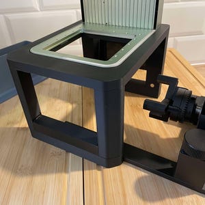 May include: A black, rectangular device with a silver interior and a green-tinted top. The device sits on a black frame with a side-mounted, black cylindrical component. The device is placed on a wooden surface.