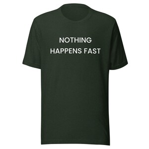 Può includere: Una t-shirt a maniche corte color verde bosco scuro con la scritta "NOTHING HAPPENS FAST" stampata in lettere maiuscole bianche sul petto. Il capo presenta un classico girocollo e un orlo dritto, offrendo un aspetto semplice e pulito.