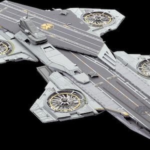 May include: A grey model aircraft carrier featuring four large circular turbine fans with gold accents. The deck displays a central runway with yellow markings and a circular emblem. The structure includes a raised command tower and detailed paneling.