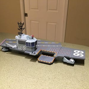 Pode incluir: Um modelo de porta-aviões cinzento com o número 99 impresso no convés e na superestrutura. O navio inclui uma torre de comando elevada, um mastro de radar, uma plataforma de elevador lateral e marcações de pista pintadas no convés.