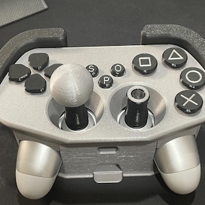 May include: A silver and black gaming controller with a joystick and various buttons, including a square, triangle, circle, and cross. The controller has a unique design with ergonomic grips and a central joystick.