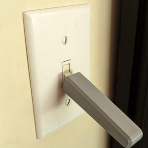 May include: A gray light switch extender attached to a white light switch plate on a light yellow wall. The extender is rectangular and angled, designed to make the switch easier to use.