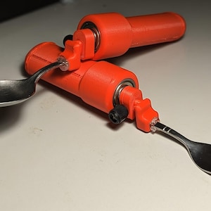 May include: Two adaptive eating utensils: a spoon and a fork. Each utensil is attached to a bright orange handle with a pivoting mechanism. The spoon and fork are made of stainless steel.