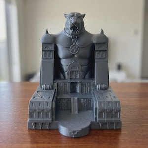 May include: A dark gray 3D printed sculpture of a feline-headed figure standing behind a fortress-like structure. The figure wears a circular pendant featuring a stylized cat emblem. The base includes architectural details like stairs and pillars.