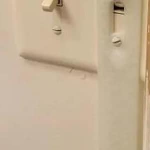 May include: A close-up of a light switch and a light switch guard. The light switch is in the off position. The guard is white and attached to the wall with screws. The wall is painted a light beige colour.