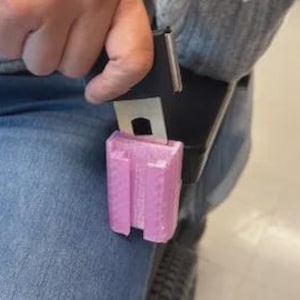 Puede incluir: Un componente de hebilla de cinturón de seguridad de metal negro con un accesorio rectangular de plástico rosa de superficie texturizada. La pieza rosa está insertada en el mecanismo de la hebilla metálica, que tiene un botón de liberación plateado.