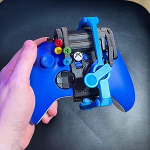 May include: A blue Xbox controller with a black and blue attachment. The controller features coloured buttons and a black directional pad. The attachment looks 3D printed and has a rotating arm.