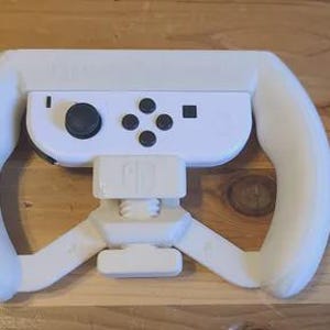 May include: A white plastic steering wheel accessory for a game controller, featuring a central mount that holds a white game controller with a black joystick and four black circular buttons, designed for a handheld gaming console experience.